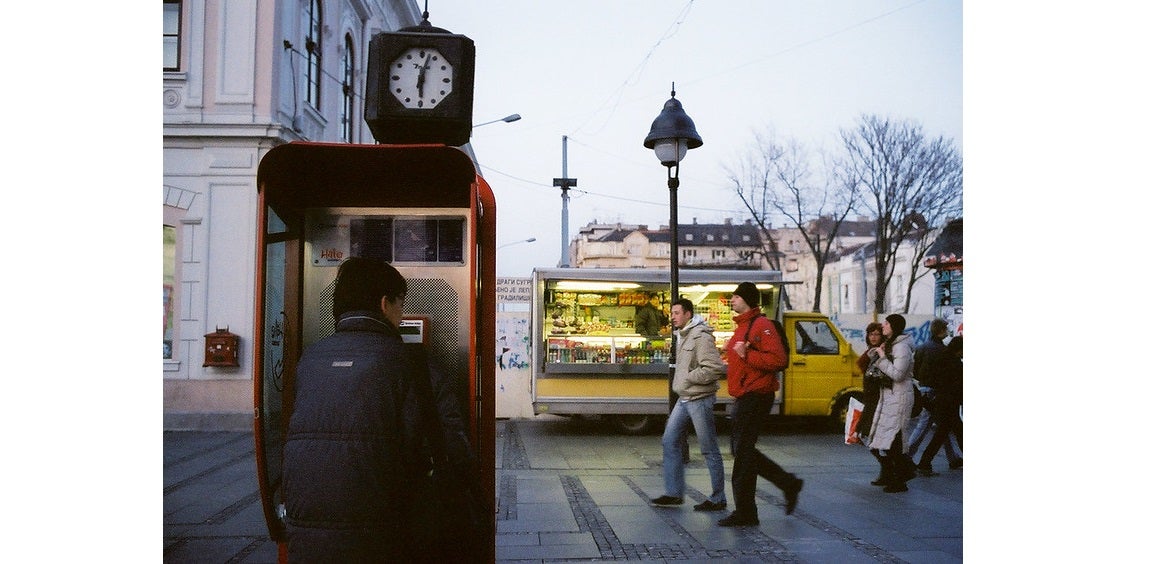 A person in winter clothes uses a public phone on a street at dusk A person in winter clothes uses a public phone on a street at dusk