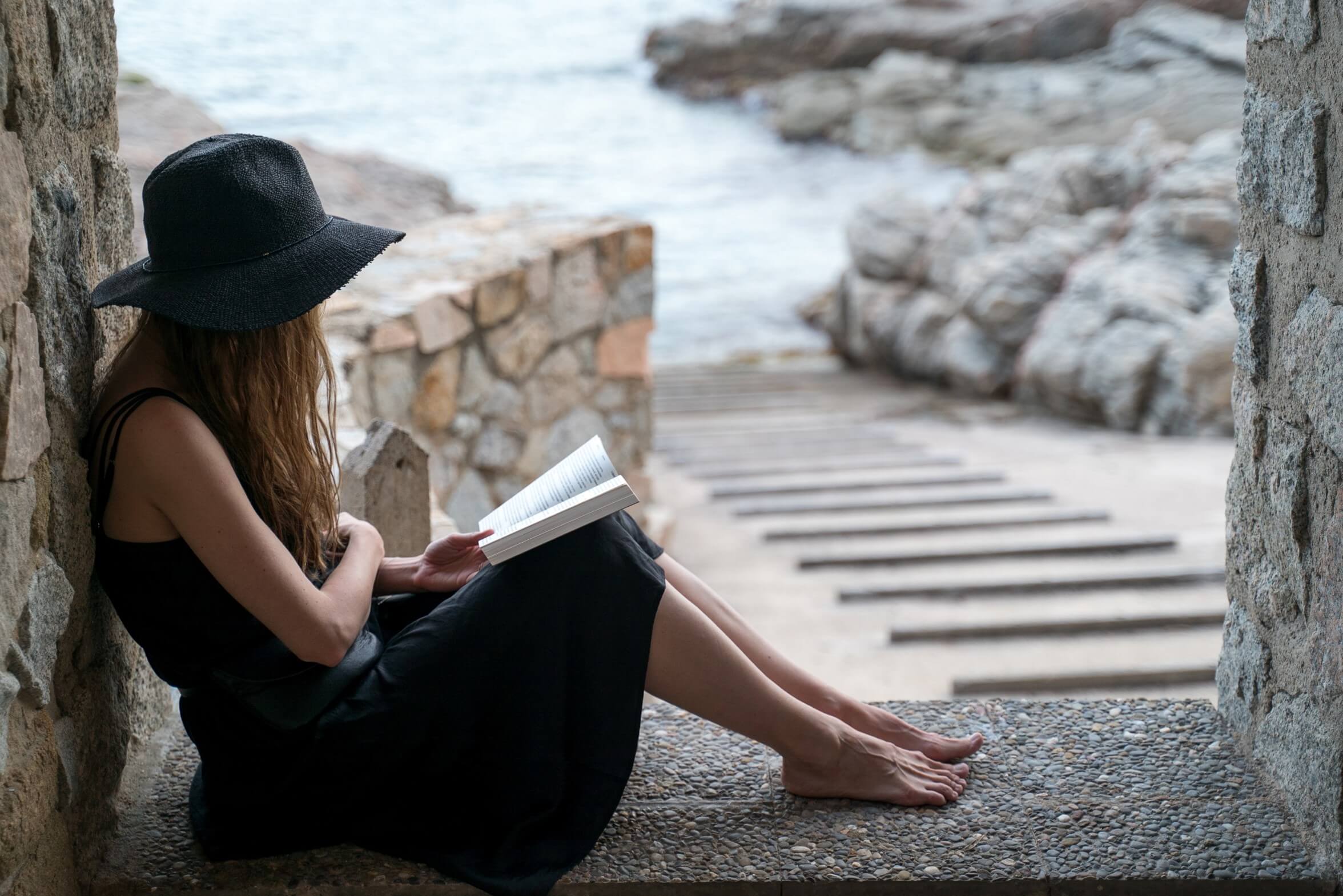 A girl in a black hat and black silk dress sits in the niche of a fishing hut on the seashore and reads a book on smart public private partnerships