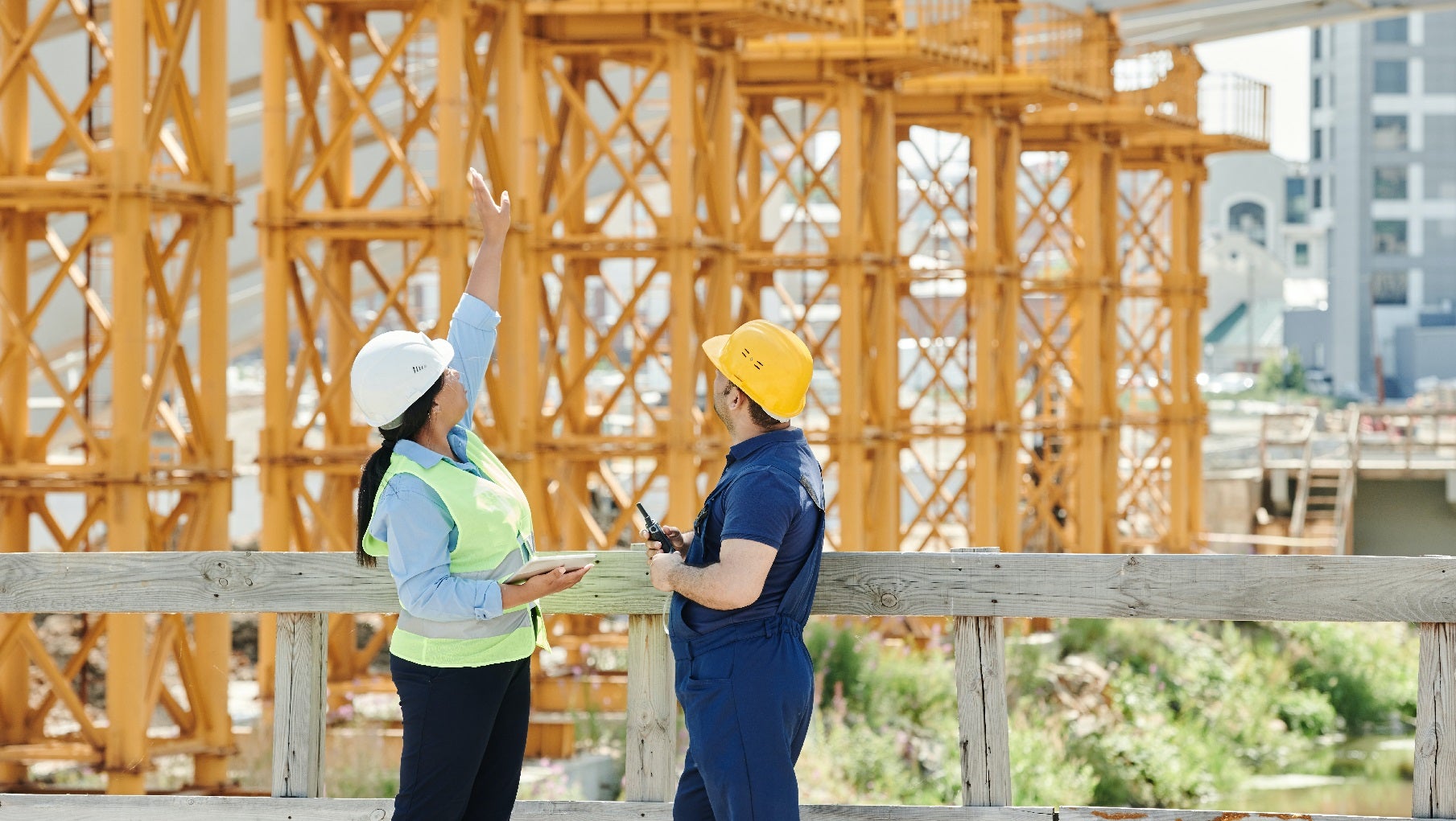 Woman and man in construction hats standing face to face, looking and pointing at construction in the background
